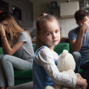 A child sits on a sofa while her mother and father sit in the background, representing child custody. - Michael Cochran Law Offices.